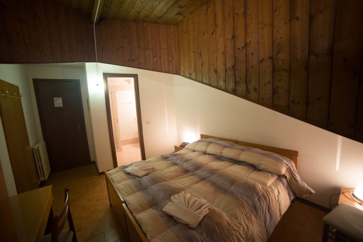 Ein Schlafzimmer mit Holzboden, einem Bett mit einem beigegrau gestreiften Bettbezug, einem Nachttisch aus Holz mit einer Lampe und einer Tür zu einem anderen Raum.