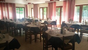 Une salle à manger avec plusieurs tables dressées, de grandes fenêtres avec des rideaux et une vue sur la verdure environnante.
