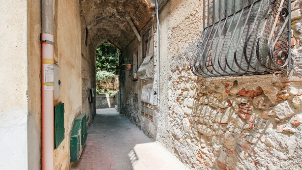 Une ruelle étroite aux murs de pierre et à la grille métallique de fenêtre, qui mène à l’entrée d’un immeuble.