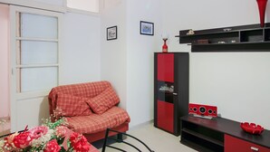 Une pièce avec un canapé rouge, un meuble noir et rouge, une petite table sur laquelle se trouve un vase rouge, et une fenêtre avec des stores.