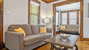Ein Wohnzimmer mit grauem Sofa, einem Holztisch und einem gemusterten Sofa im Hintergrund.