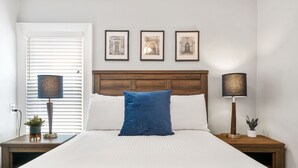 Ein ordentlich gemachtes Bett mit einem blauen Kopfkissen, zwei Nachttischlampen und drei gerahmten Bildern an der Wand.