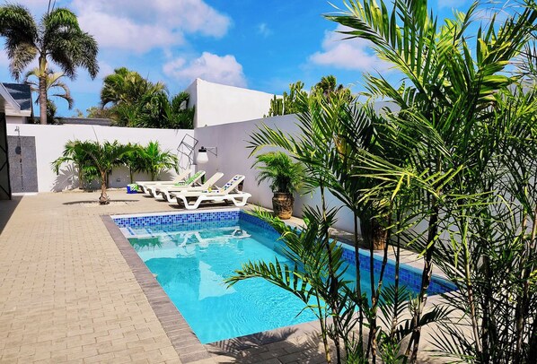 Piscina com espreguiçadeiras, cercada por plantas e uma parede branca.