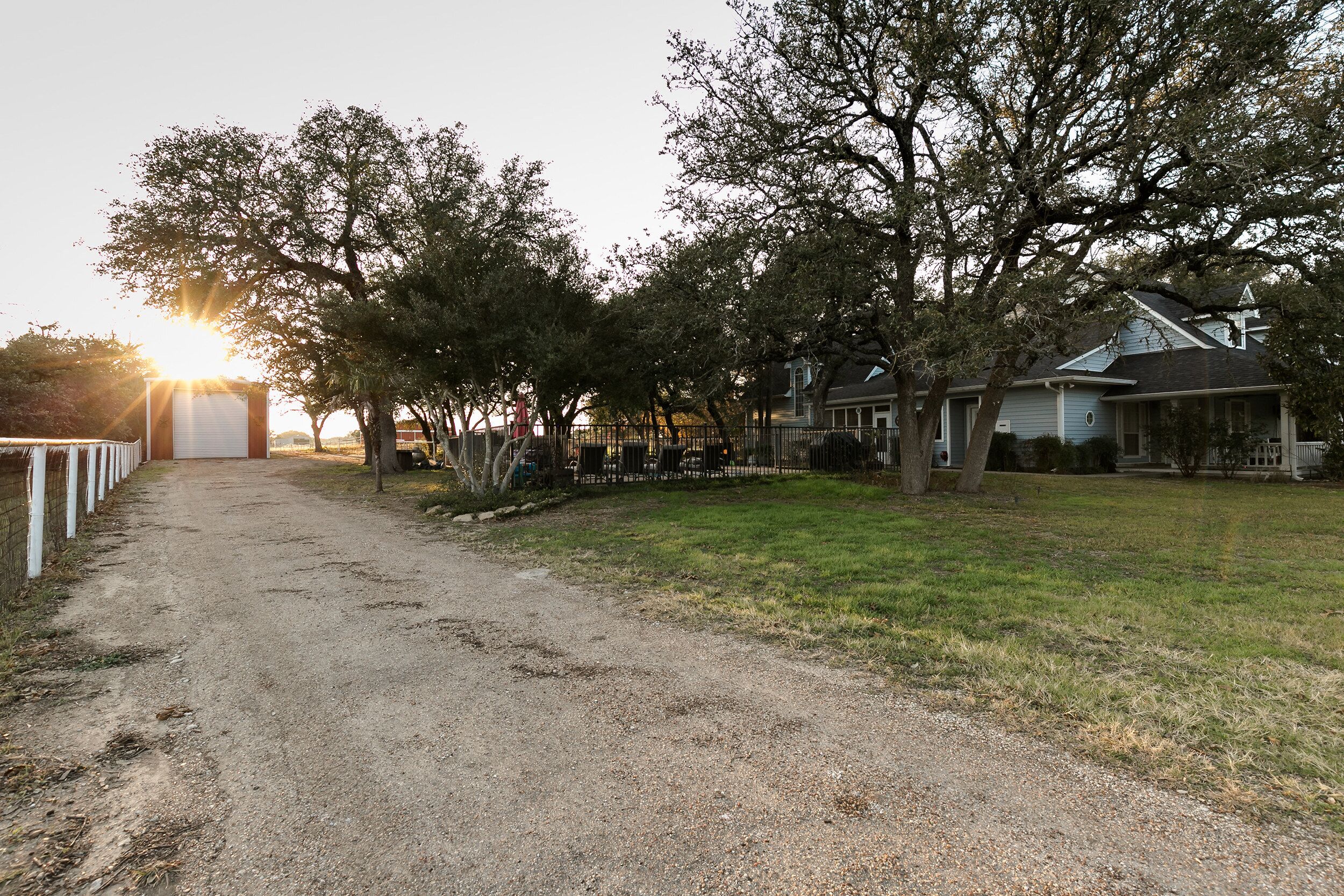 A rural road leading to a row of houses with a fence on one side and trees lining the street.