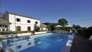 Una piscina rectangular con agua azul cristalina, rodeada por un patio de ladrillo. Se observan sillones de descanso y sombrillas al fondo, y un edificio blanco con contraventanas marrones es visible.