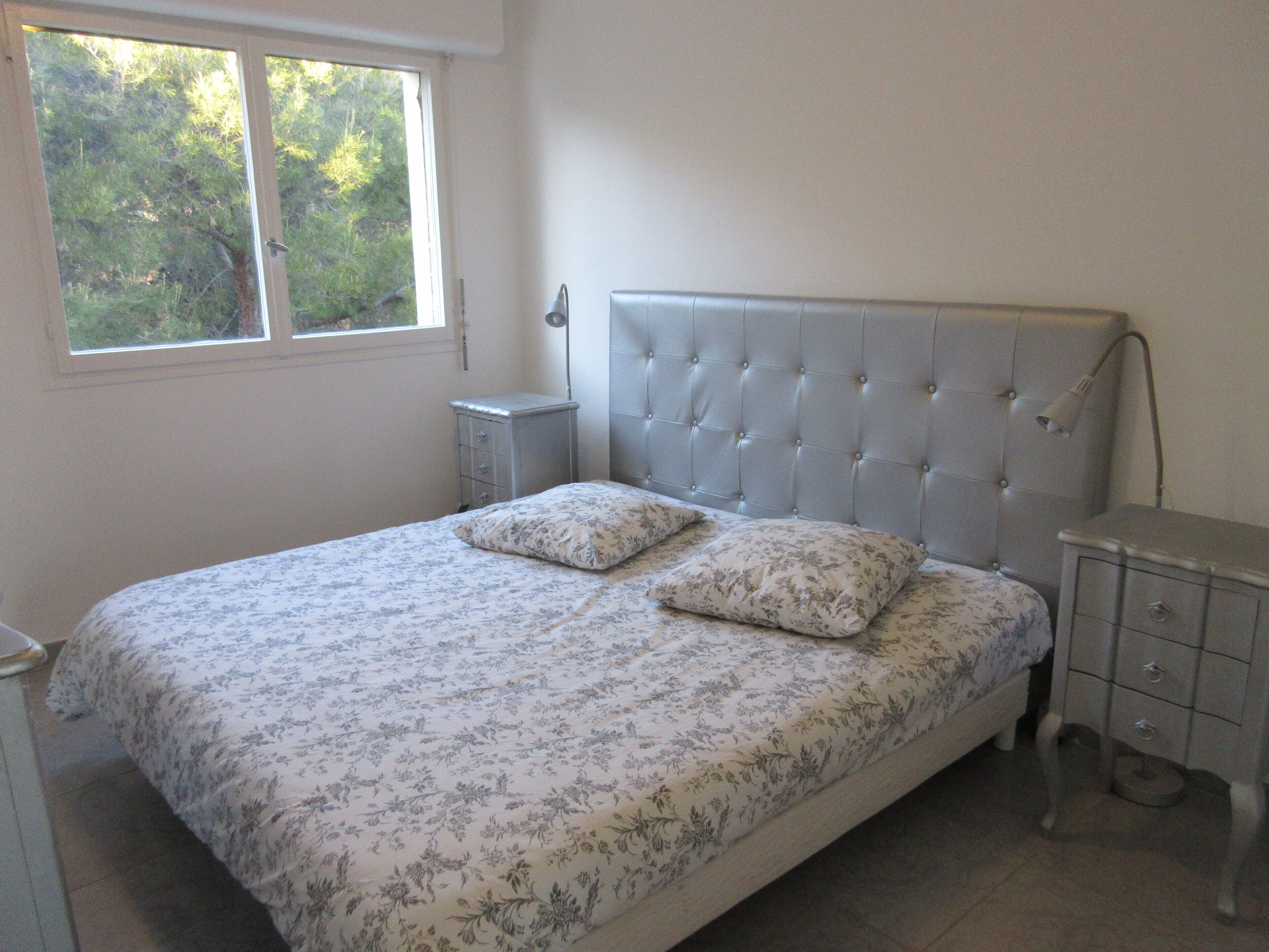 Une chambre à coucher avec un lit, deux oreillers, une table de chevet, une lampe et une fenêtre donnant sur des arbres.