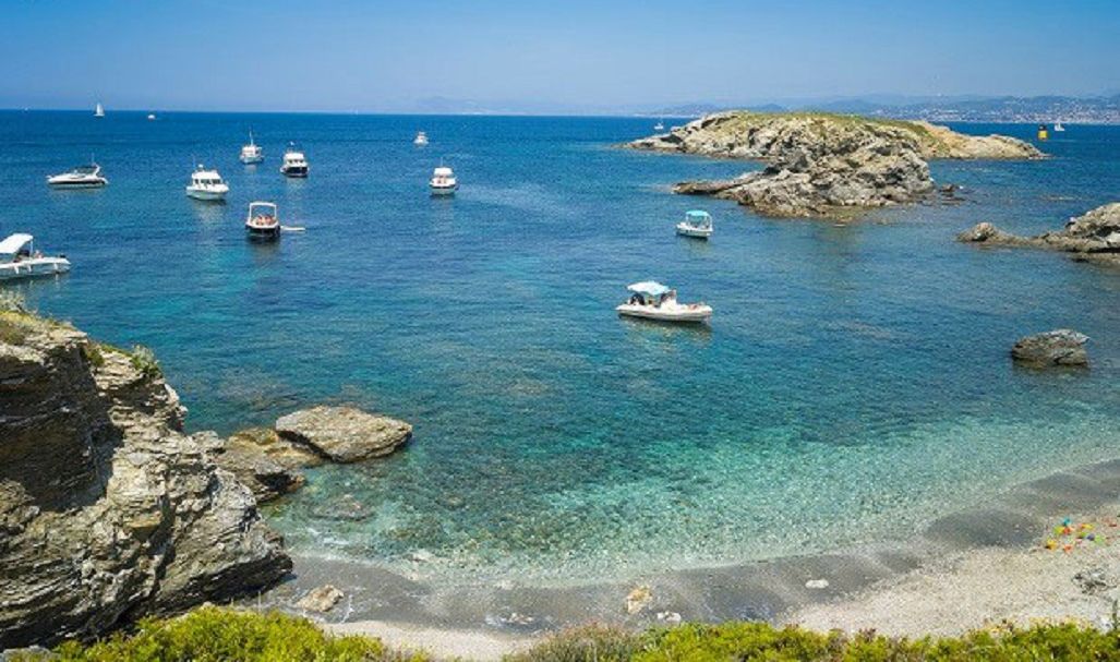 A coastal scene with boats on clear blue water, rocky outcrops, and a distant cityscape.