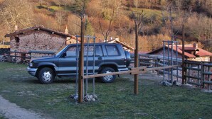 Une cabane rustique aux murs en pierre et au toit recouvert de tuiles rouges, entourée d’une clôture en bois et avec un VUS garé à proximité.
