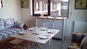 A cozy dining area with a wooden table set for four, a grey sofa with patterned cushions, and a window overlooking a building.