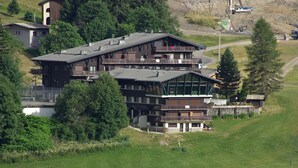 A multi-story building with balconies, surrounded by greenery and a hilly landscape.