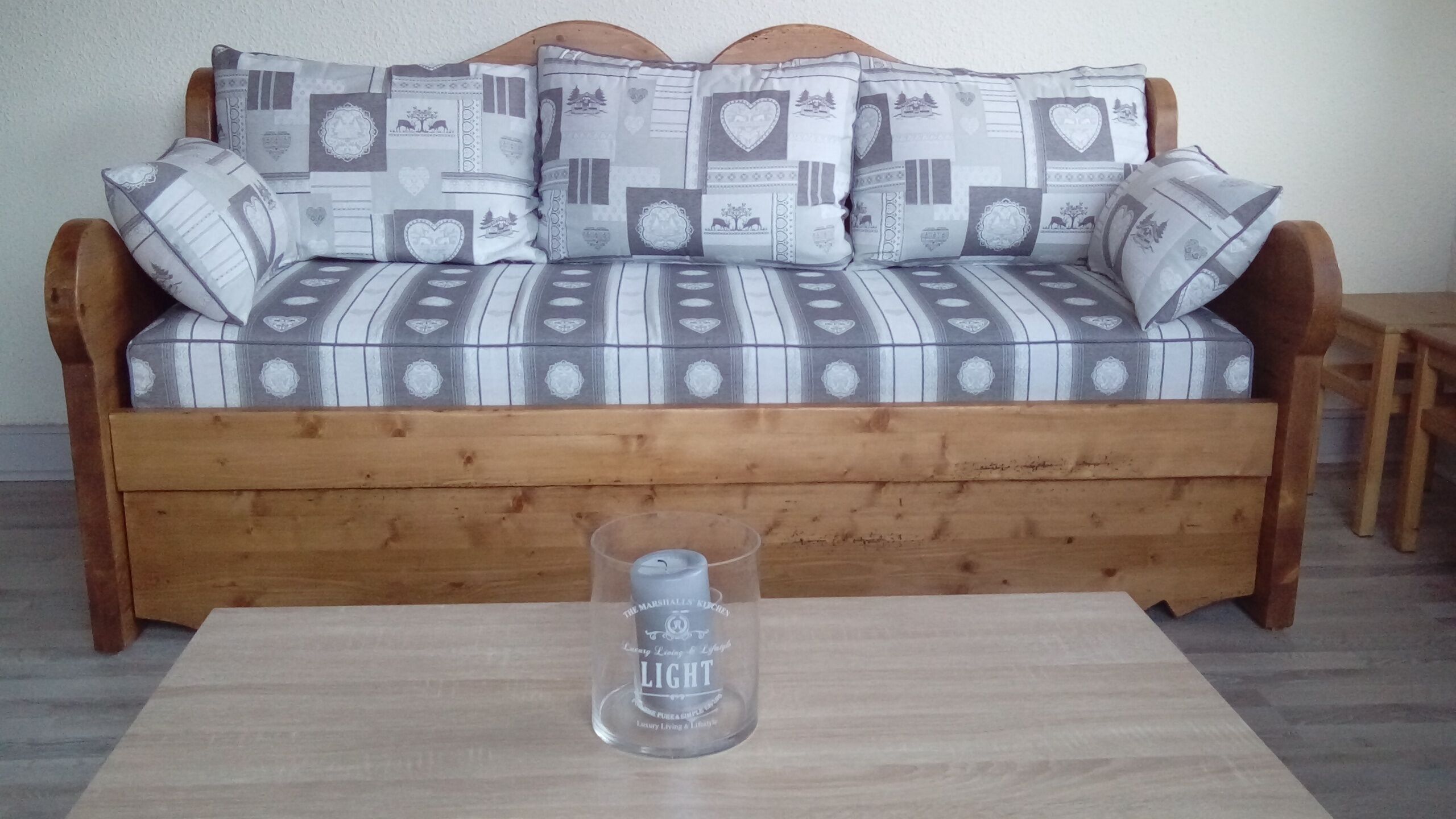 Un divano in legno con cuscini decorati, una bottiglia di vetro trasparente su un tavolino in legno e una parete liscia sullo sfondo.