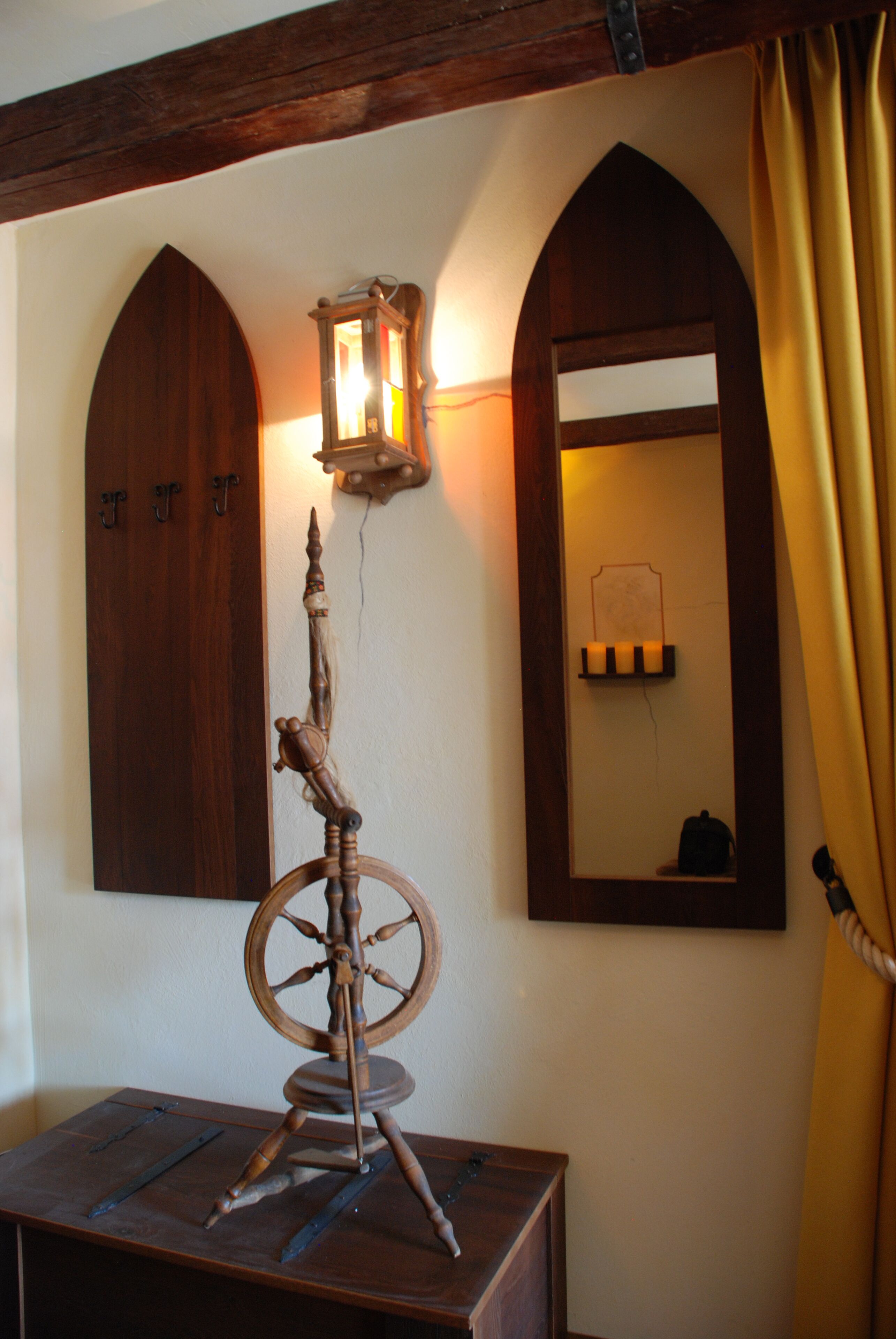 Une roue à filer en bois posée sur une table, un miroir fixé au mur et une porte en bois avec des crochets.