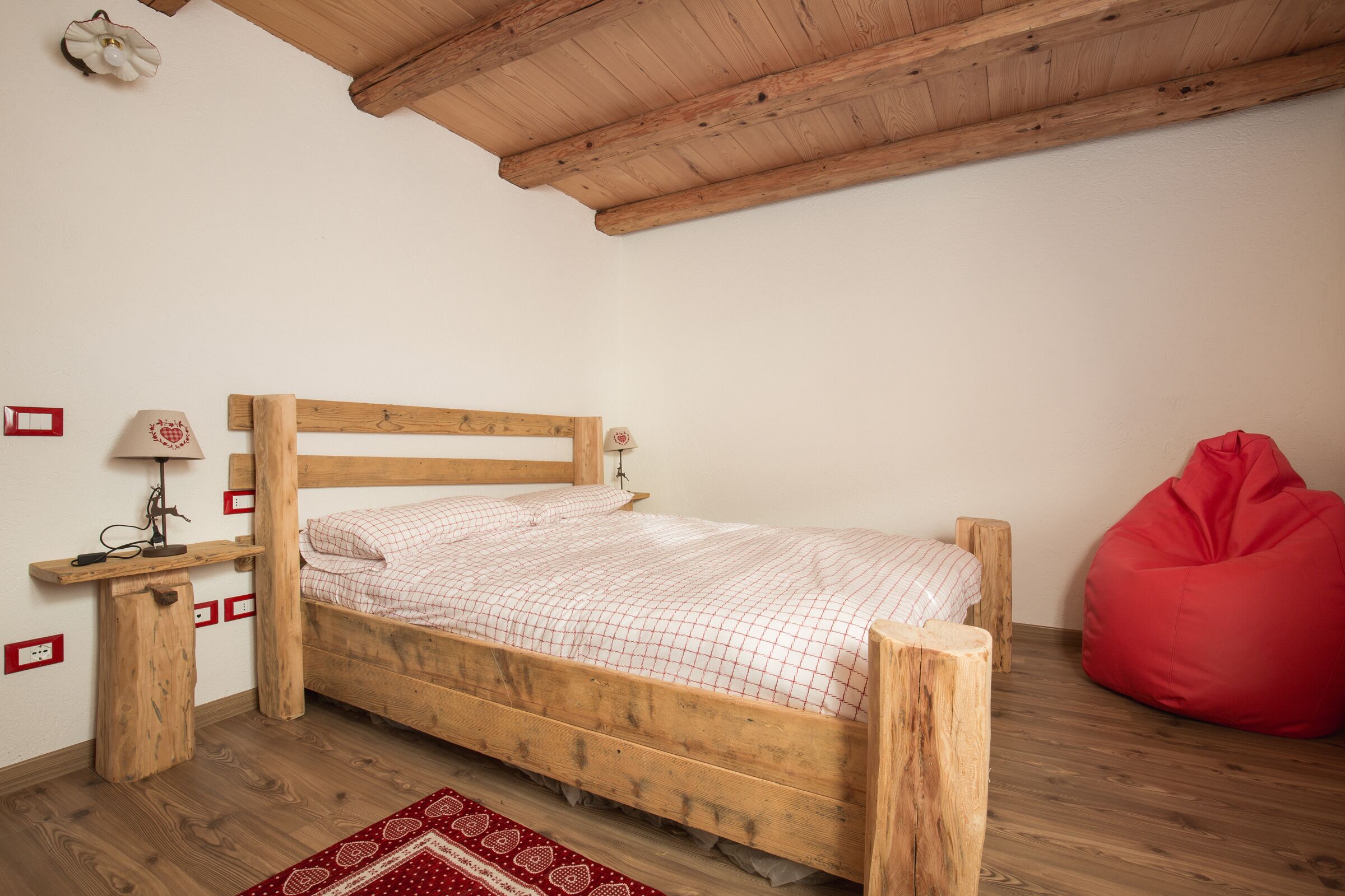 Ein hölzernes Bett mit karierter Bettdecke, ein roter Sitzsack und ein kleiner Tisch mit einer Lampe.