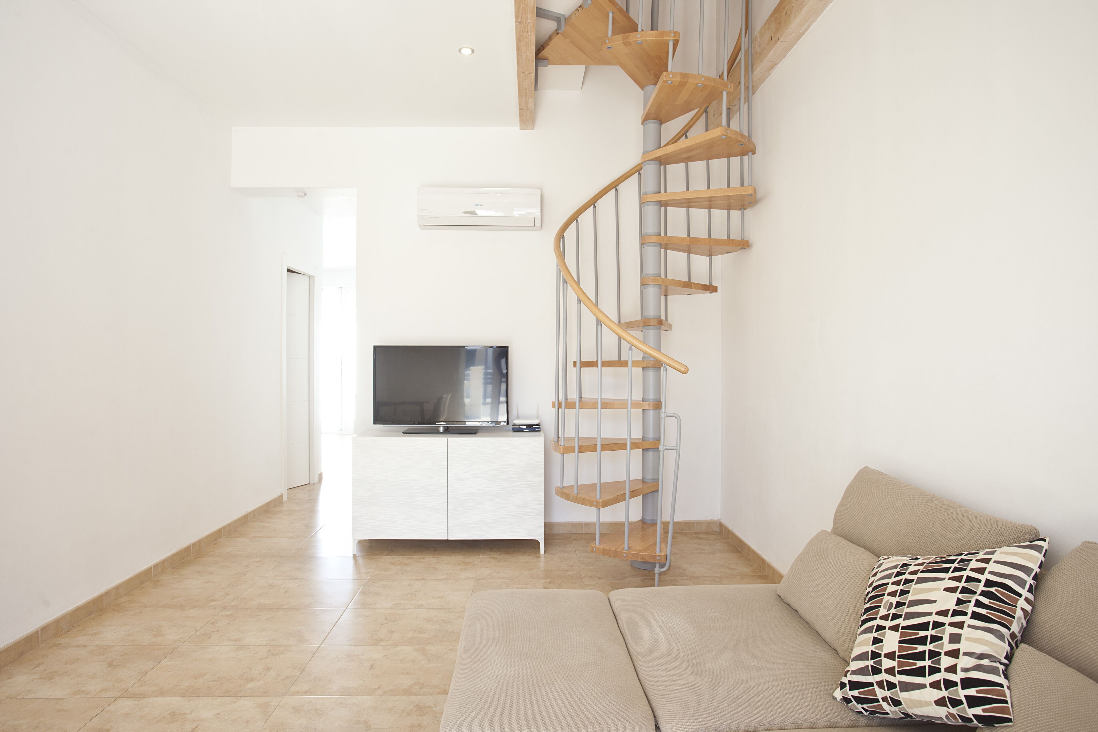 A modern living room with a spiral staircase, a flat-screen TV, a sofa, and a patterned pillow.