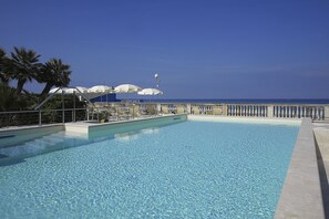 Ein klares, blaues Schwimmbecken mit Liegestühlen und Sonnenschirmen, eine Palme und Blick auf den Ozean.