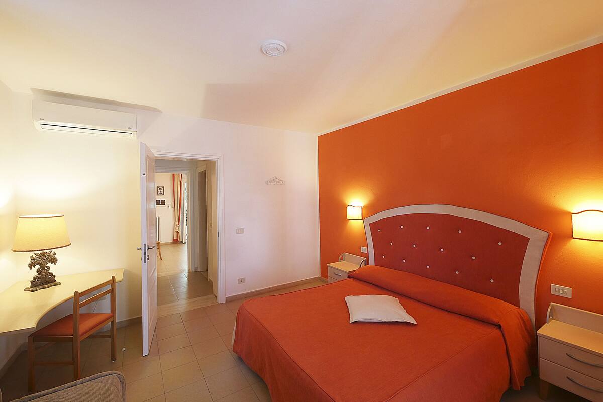A hotel room with a bed, bedside tables, a desk with a chair, and a door leading to another room.