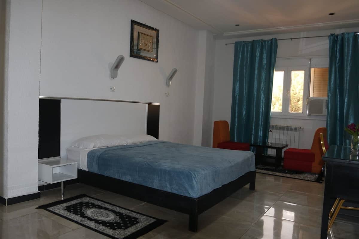 A bedroom with a bed, a nightstand, a desk, two chairs, a window with curtains, and a radiator.