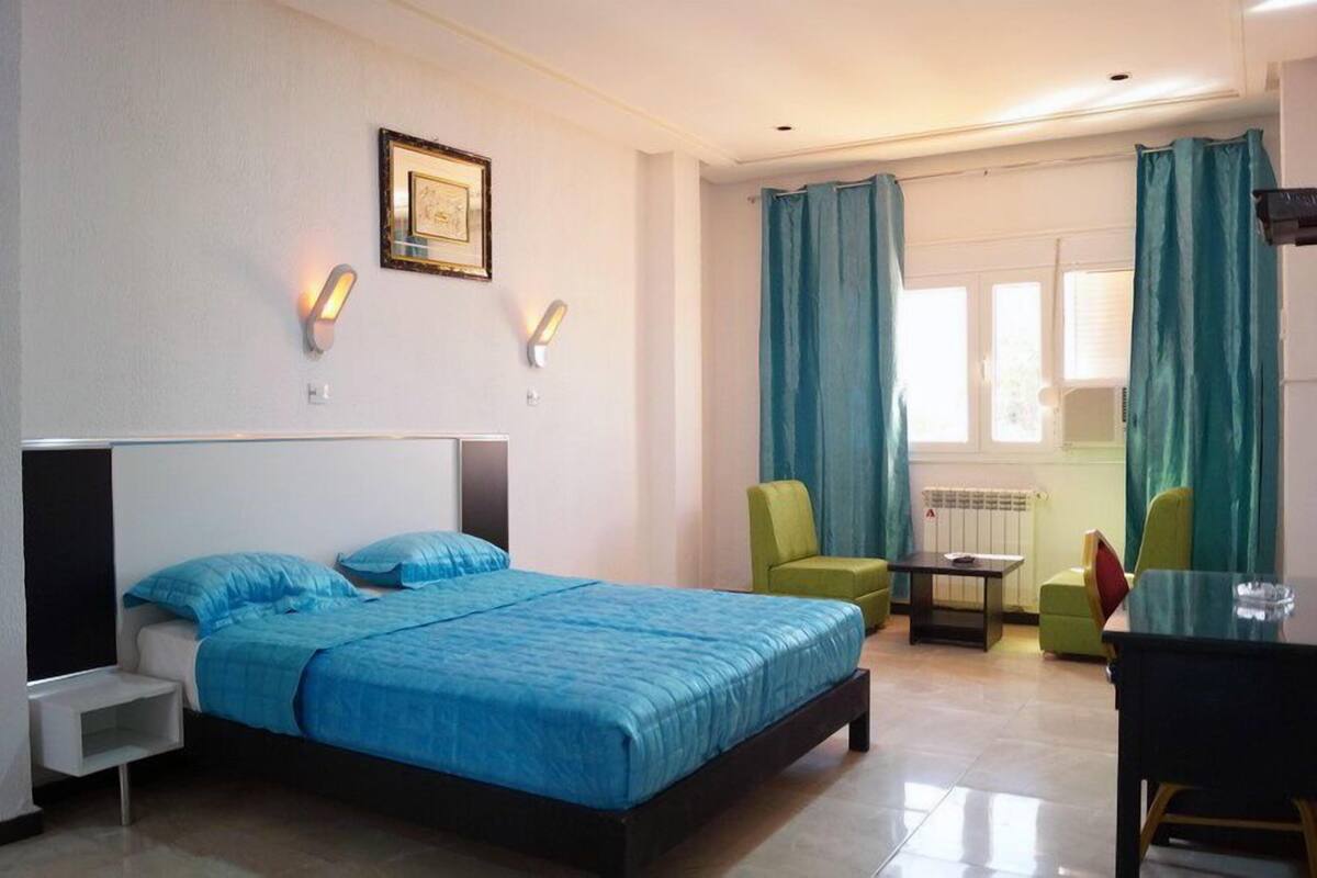 A hotel room with a bed, two chairs, a small table, and a window with curtains.