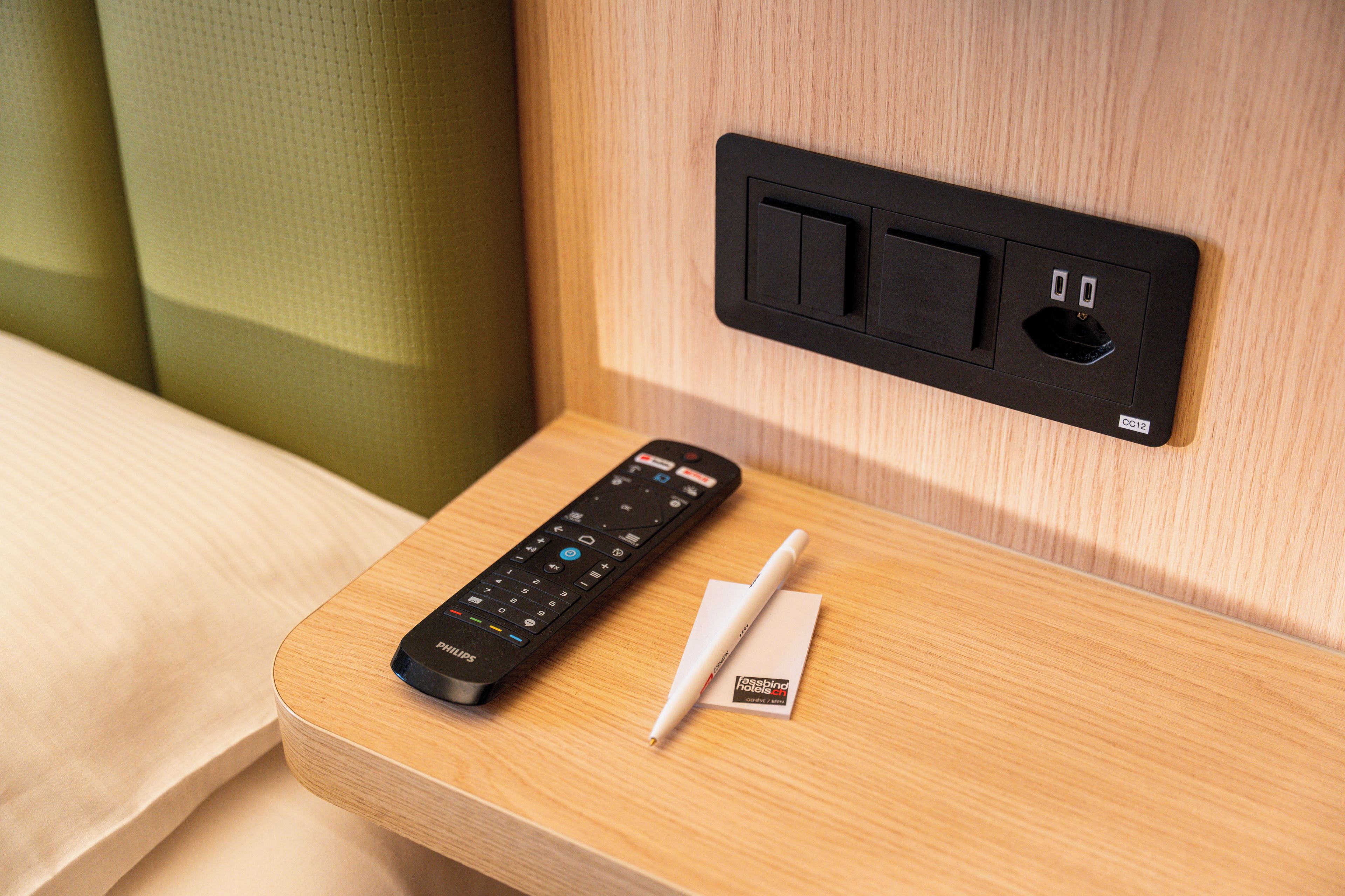 Un control remoto y un bolígrafo sobre una mesita de noche de madera con un enchufe y puertos USB.