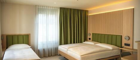 A hotel room with two beds, a desk, a chair, a window with curtains, and a wall-mounted clock.