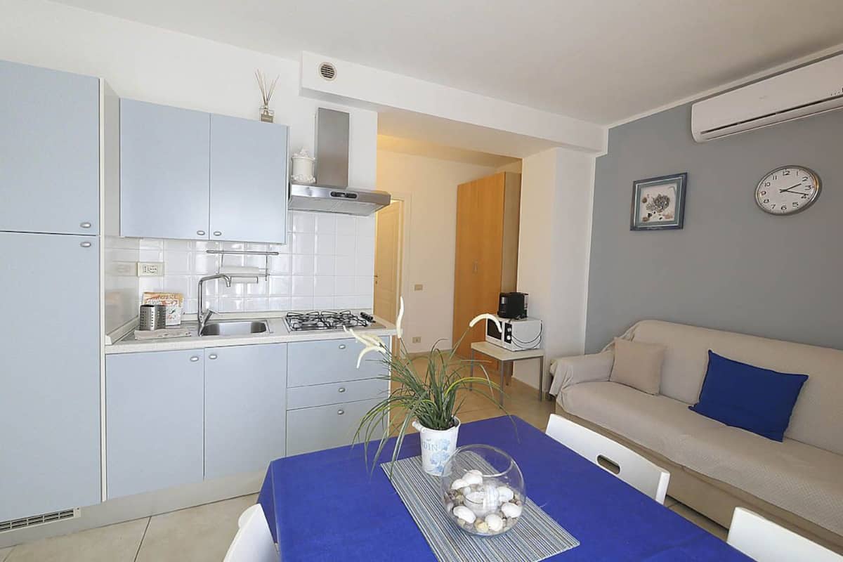 A compact kitchen with modern appliances, a dining table with a blue tablecloth, and a sofa in a living area.