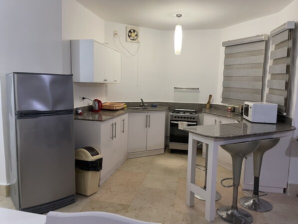 Una cocina compacta con gabinetes blancos, un refrigerador de acero inoxidable, una estufa a gas, un microondas y un pequeño bote de basura.