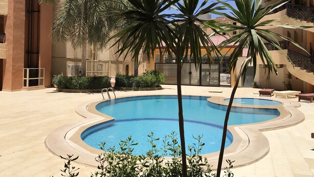 A swimming pool with a curved design, surrounded by palm trees and a building with balconies. - One bedroom (403) touristic area, near to Mamsha Street (Hurghada)
