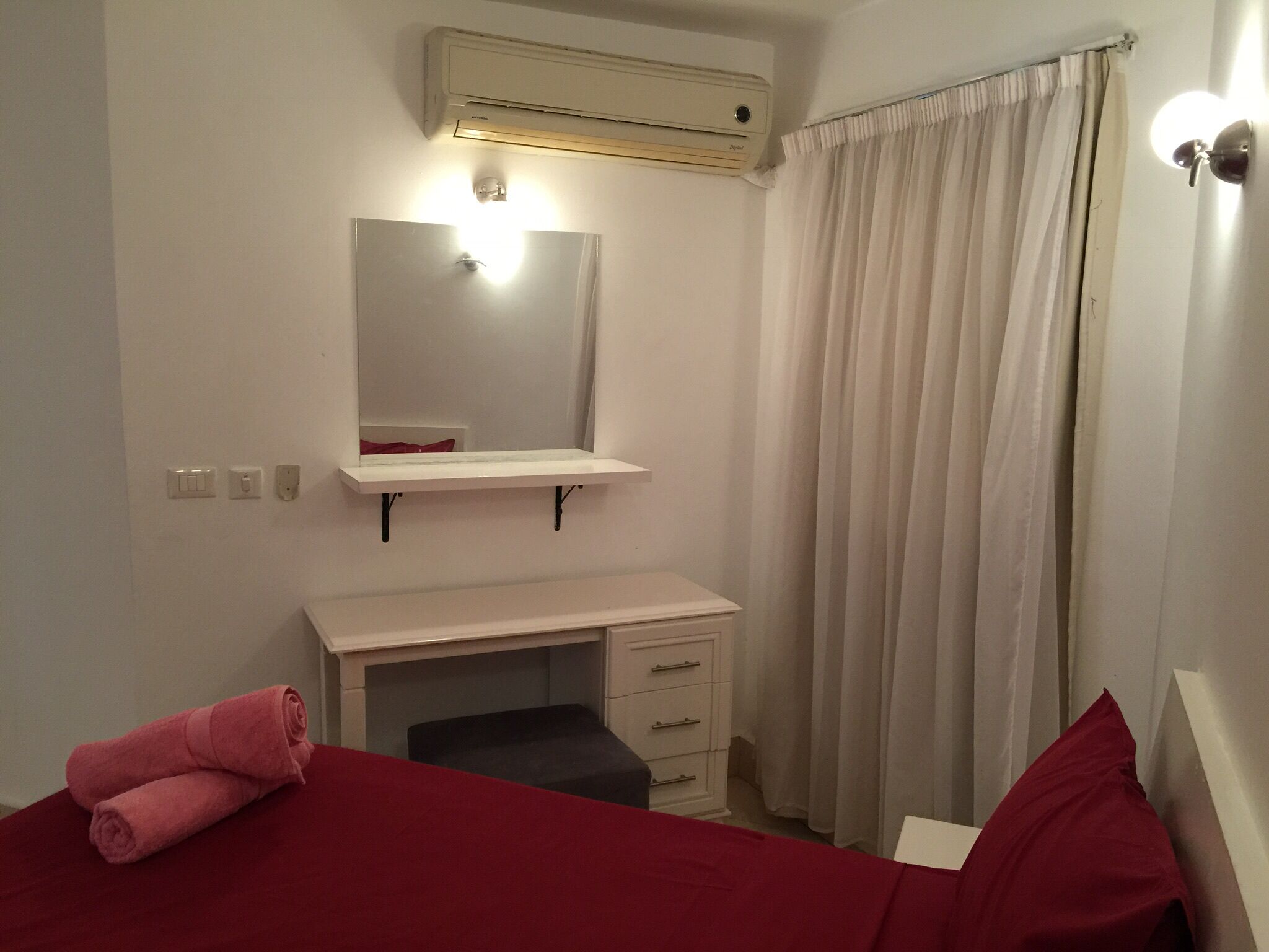 Un dormitorio con cama, un escritorio pequeño, un espejo, aire acondicionado y una ventana con cortinas.
