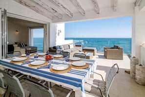 Uma área de refeições ao ar livre com vista para o mar, com uma mesa posta, incluindo pratos, copos e uma tigela de frutas vermelhas.