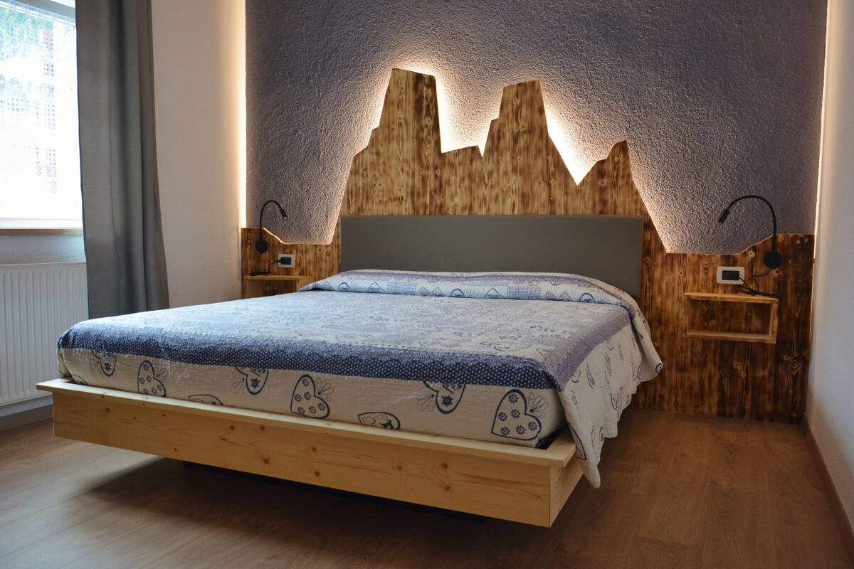 Ein Schlafzimmer mit einem Holz-Kopfteil, einem Bett mit einem gemusterten Bettbezug, Nachttischen und einem Fenster mit Vorhängen.