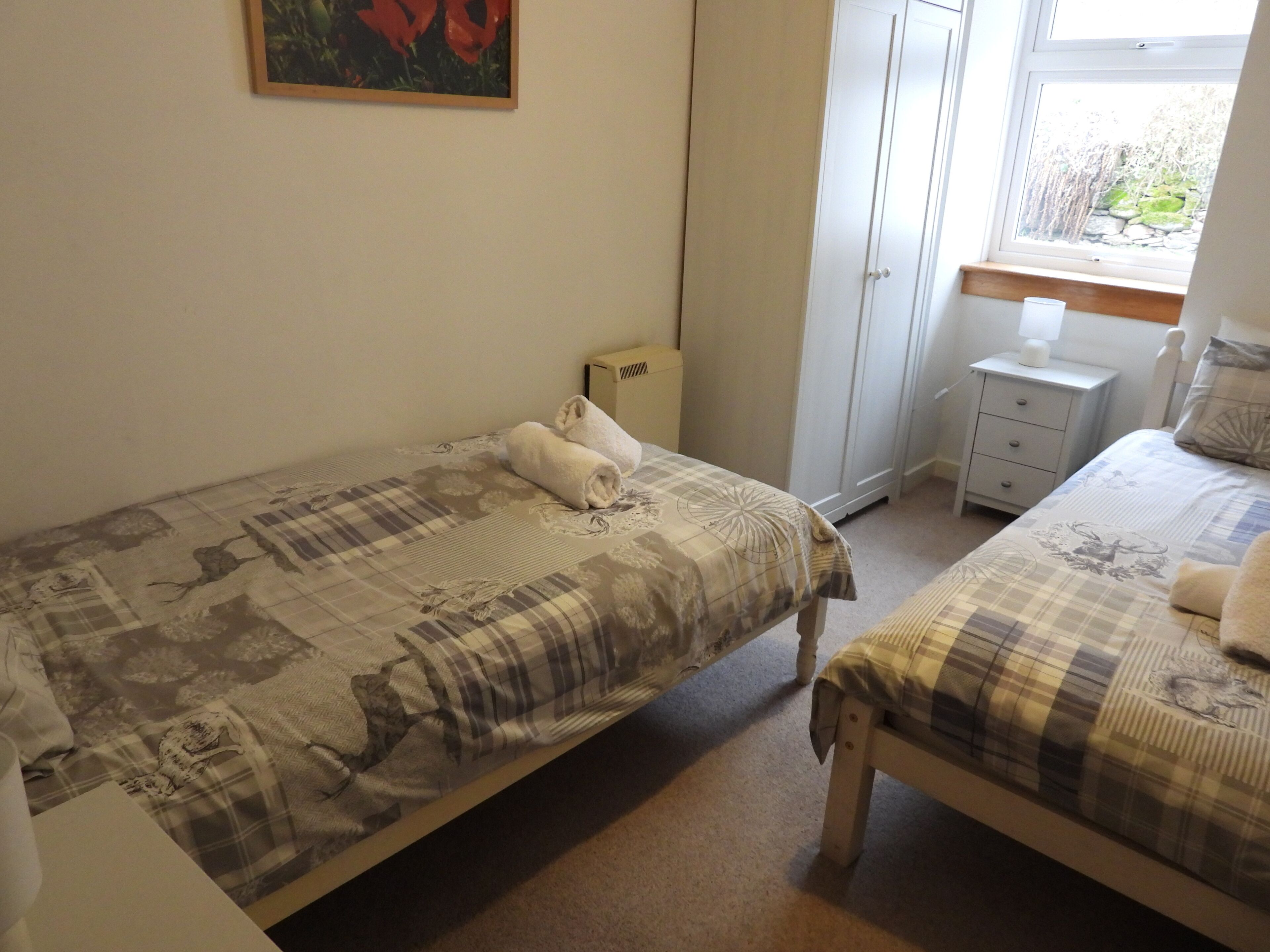 A bedroom with two beds, a wardrobe, a bedside table with a lamp, and a window with a view of greenery.