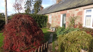 A stone house with a green door, a stone chimney, and a garden with a large bush and a hedge. - Smithy Cottage, Perthshire (Perth)