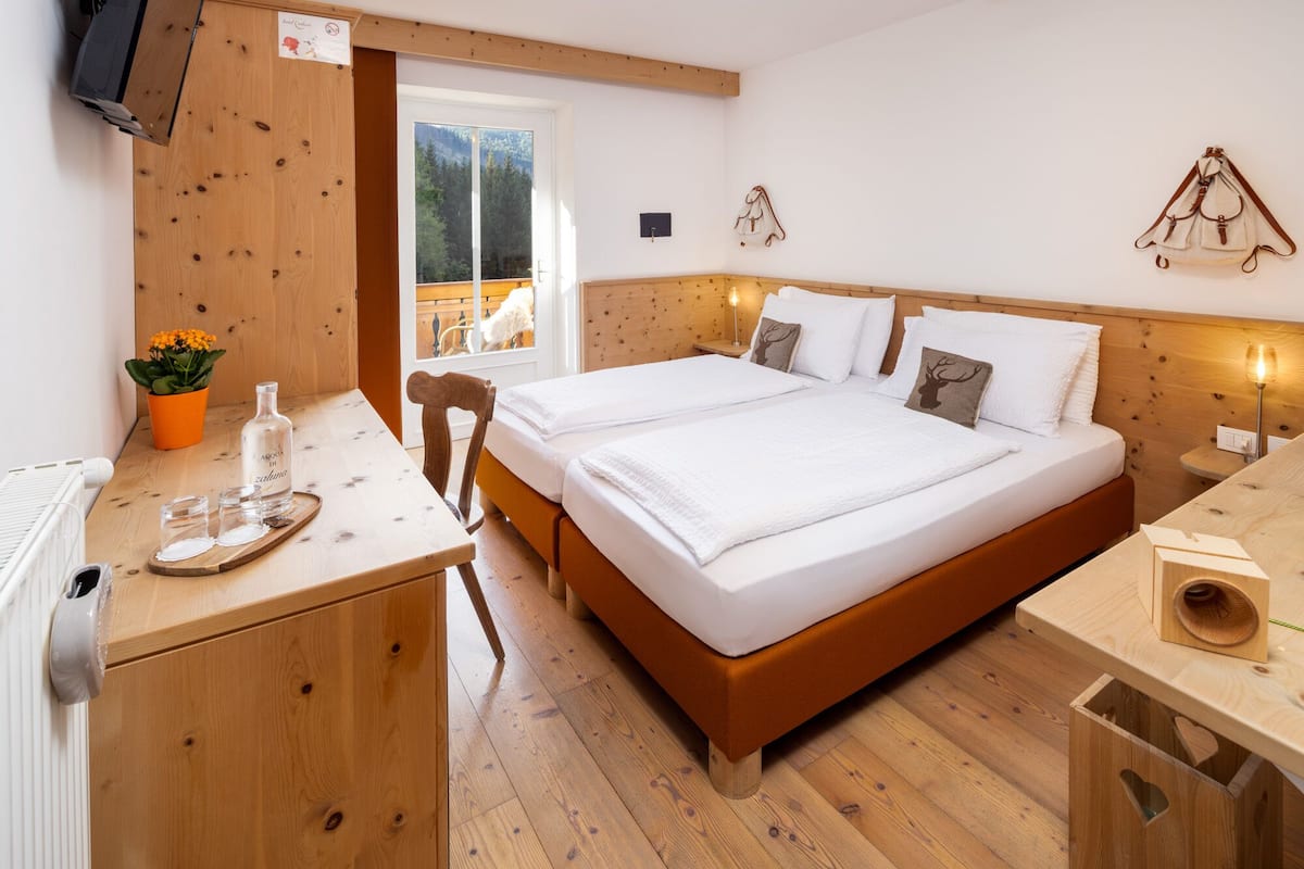 Un dormitorio de madera con una cama doble, una mesita con una botella y vasos, una planta en maceta y vistas al exterior.