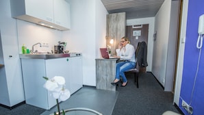 Ett modernt hotellrum med ett litet pentry, en sittgrupp med en bärbar dator och ett litet bord med en vas med blommor.
