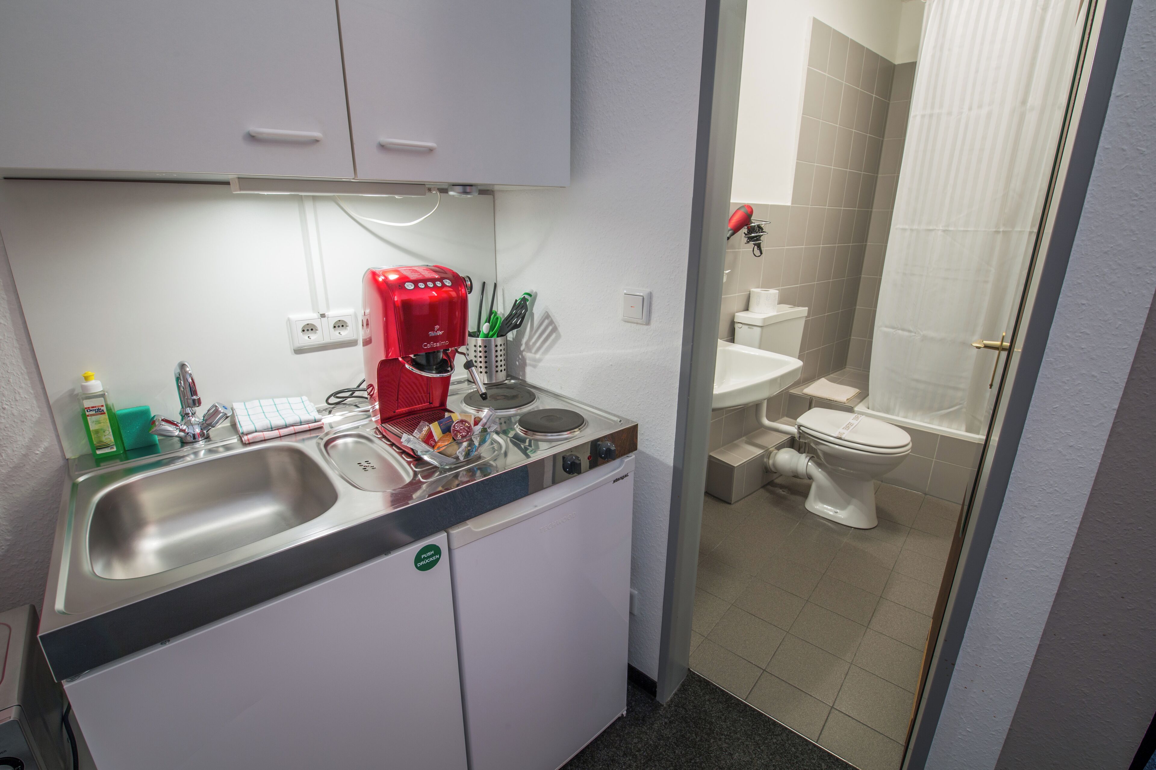 Een kleine keuken met een spoelbak, een rode koffieapparaat en een kleine koelkast. Grenzend aan de keuken bevindt zich een badkamer met toilet en douche.