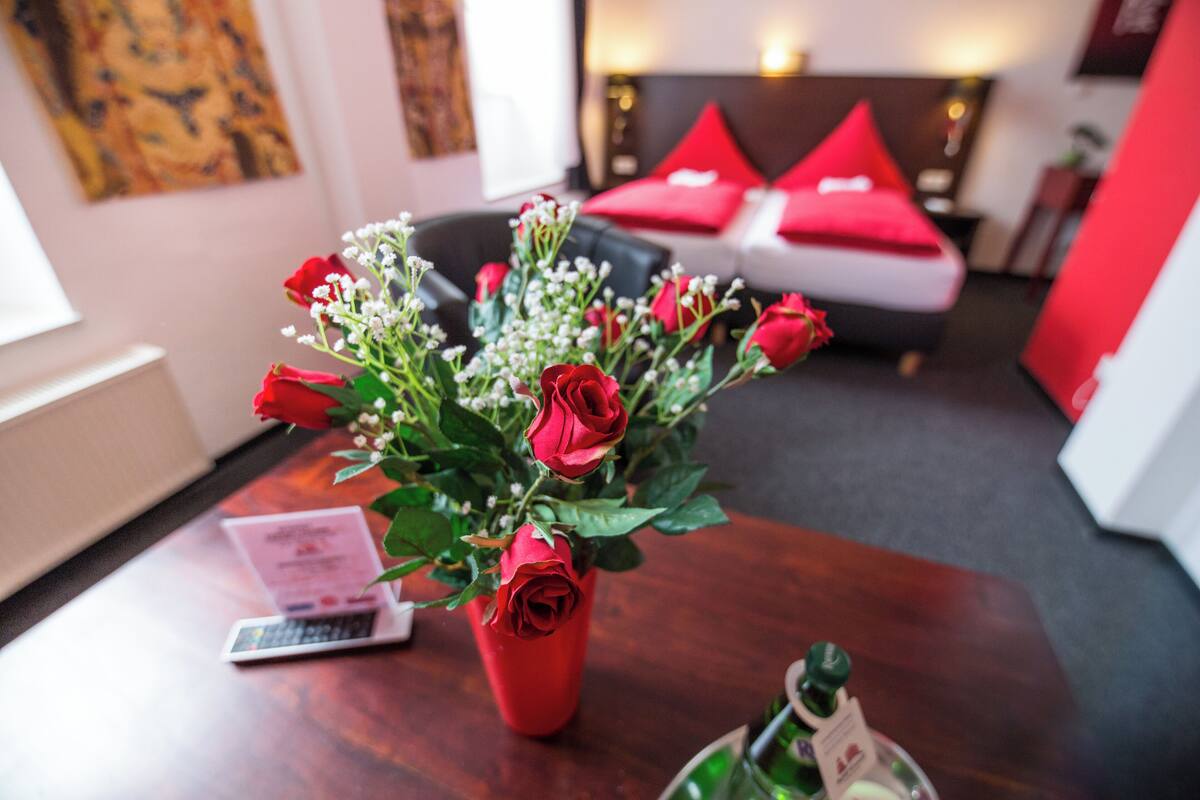 A room with a wooden table, a red vase with roses, a bottle, and a laptop.