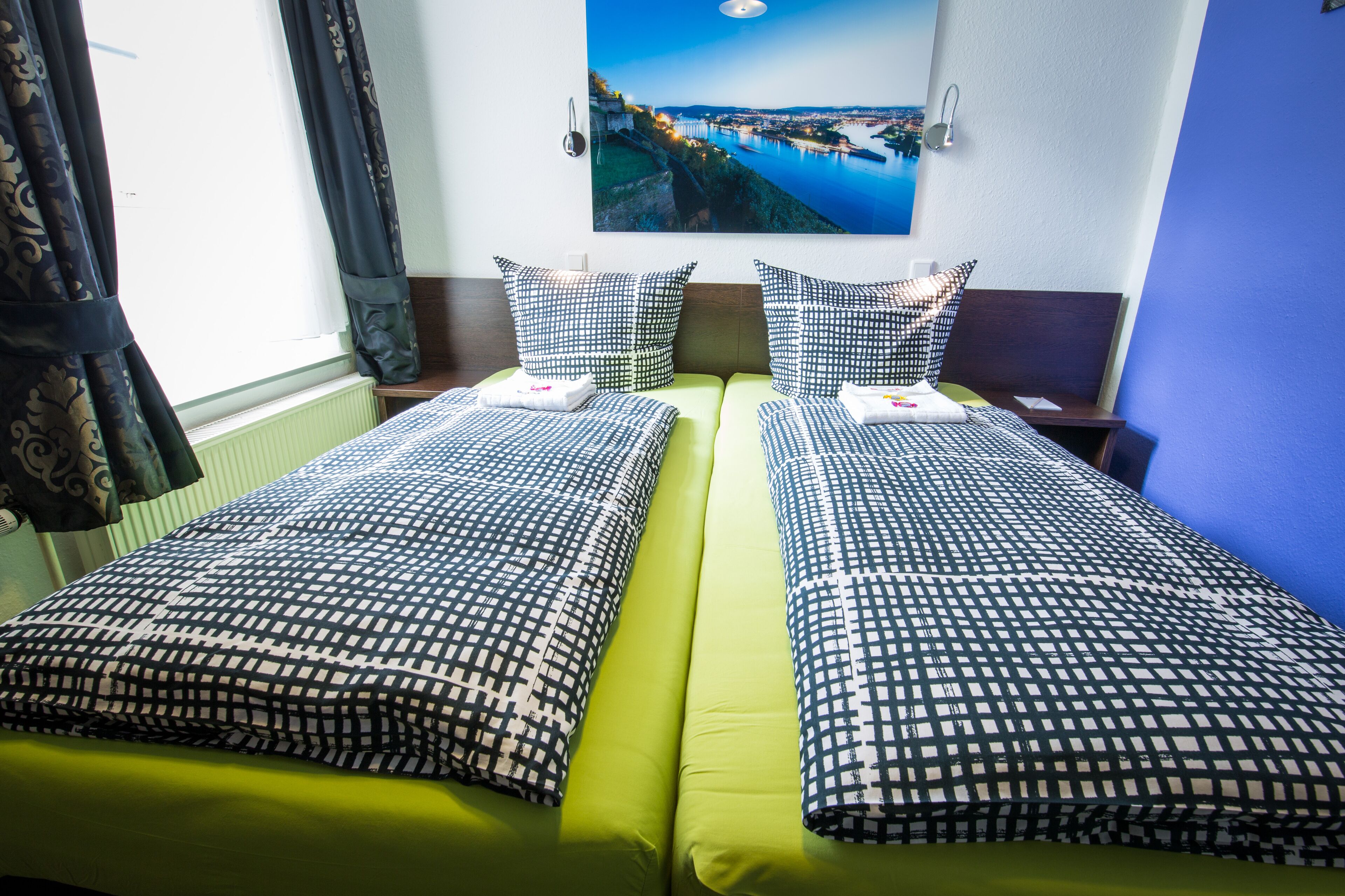 A hotel room with two beds, each with a patterned bedspread and pillows, a green mattress, and a large window with curtains.