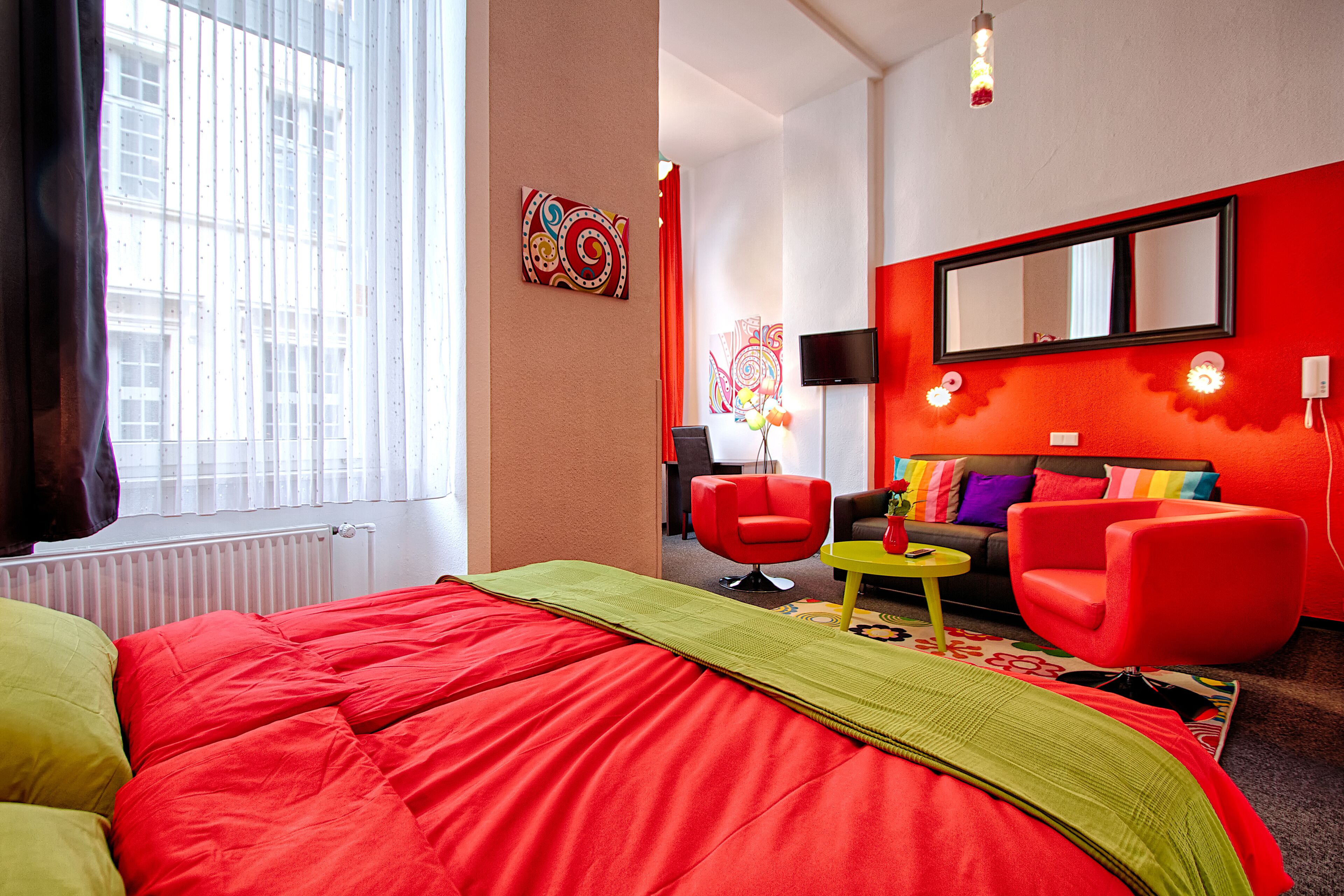 Un moderno cuarto de hotel con una cama grande, ropa de cama verde y roja, un sofá rojo, una pequeña mesa amarilla y un espejo grande en la pared.