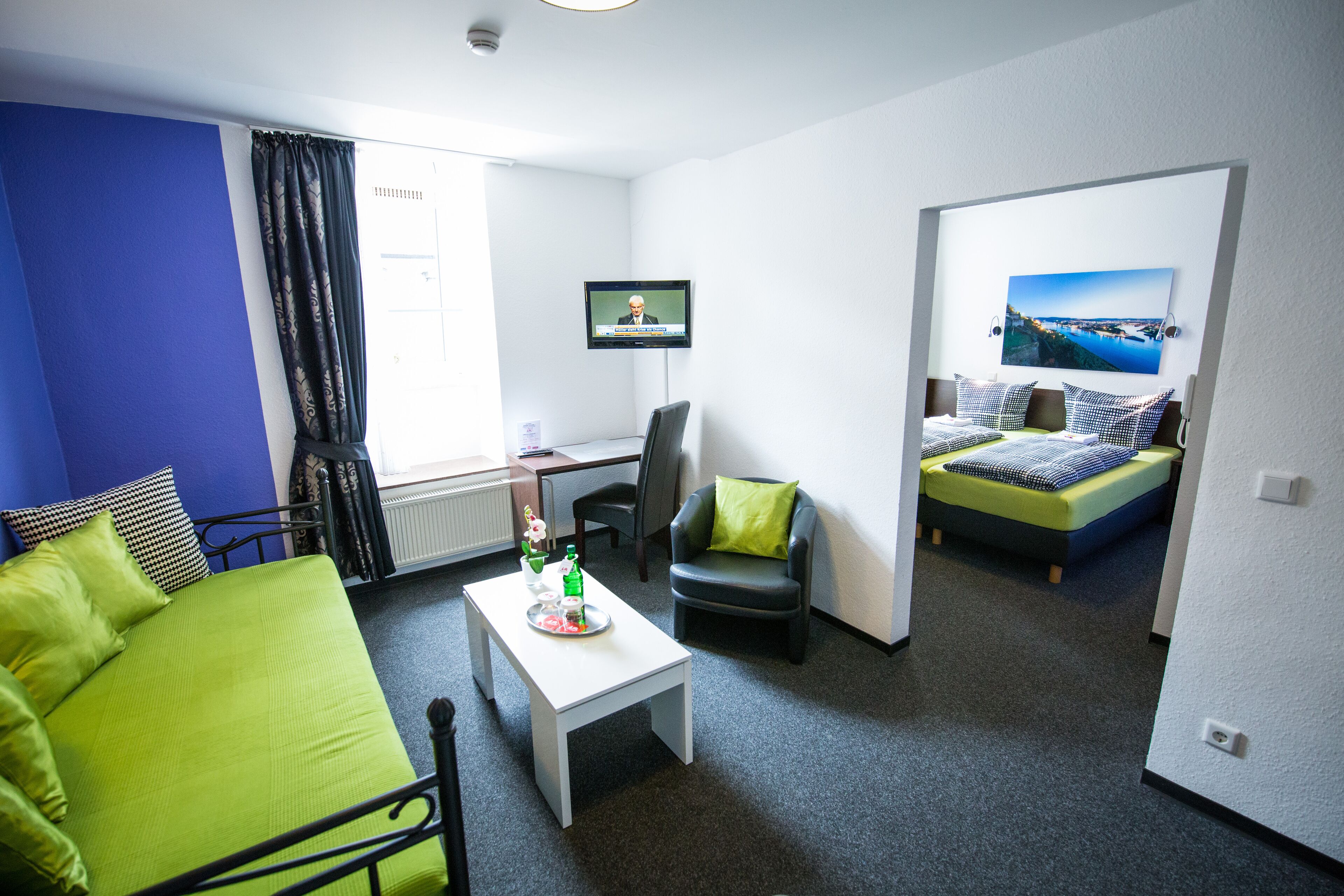 A hotel room with a green bed, a white desk, a chair, a TV, and a painting on the wall.