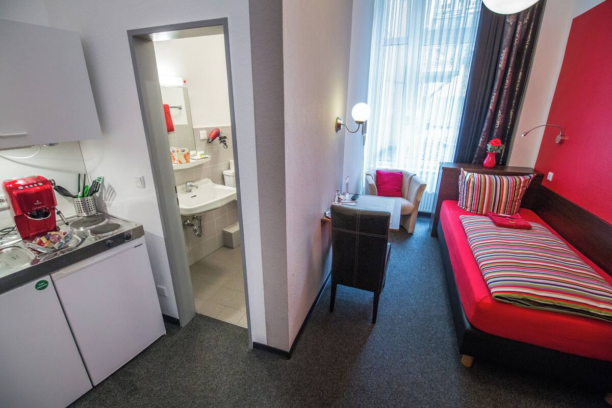 A compact hotel room with a kitchenette, a small bathroom, a bed with striped bedding, and a window with curtains.