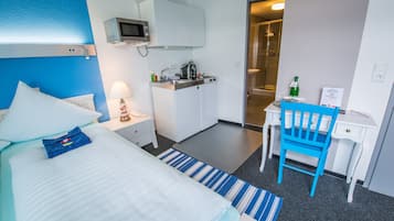 A hotel room with a bed, a desk, a chair, a kitchenette, and a shower area.