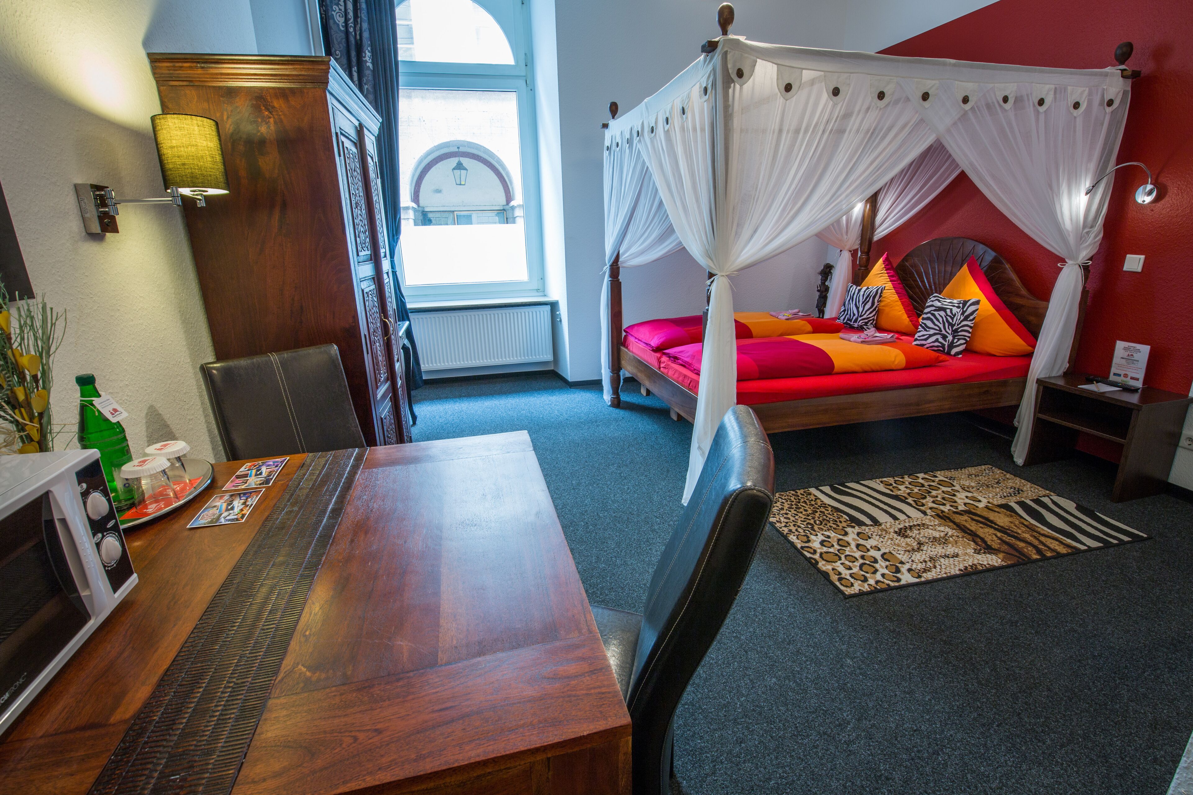 A room with a four-poster bed, a wooden table, a microwave, and a view of the street through a window.