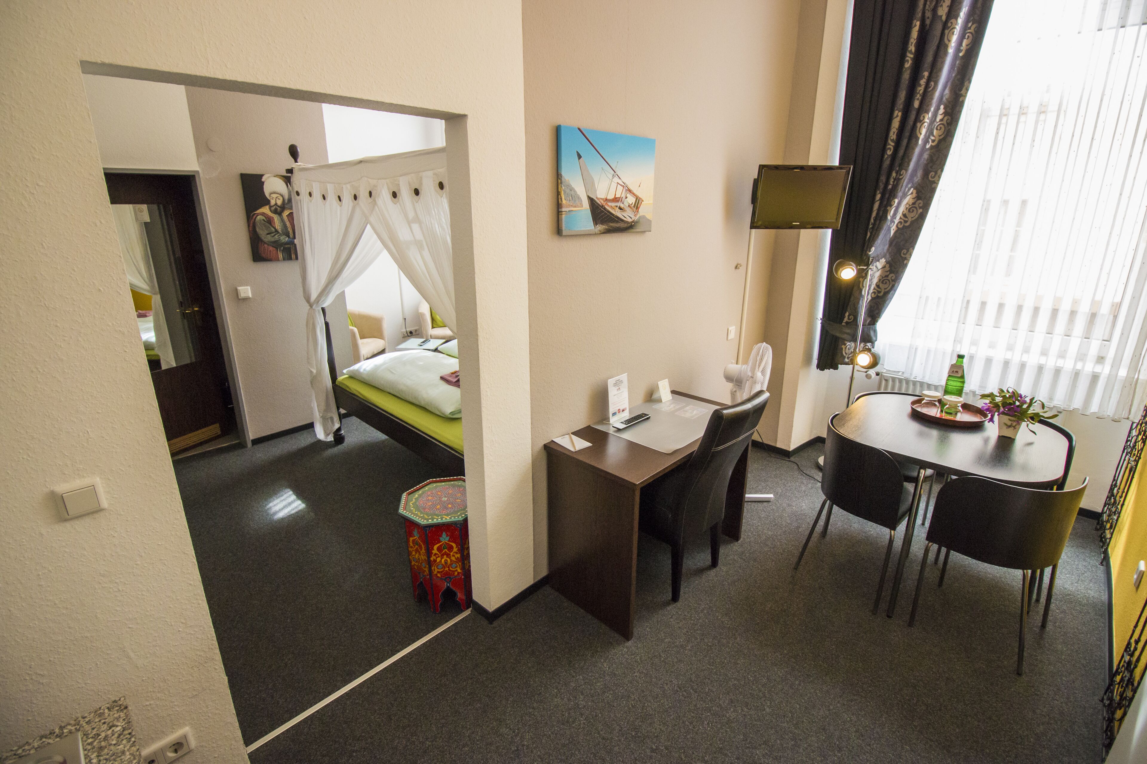 A hotel room with a bed, a desk, a chair, a small table with a plant, and a painting on the wall.