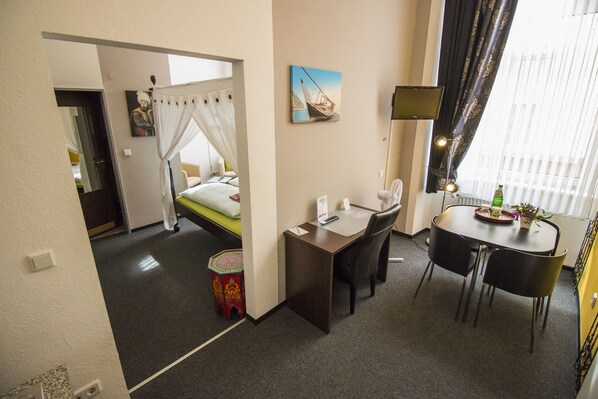 Habitación de hotel con cama, escritorio, silla, una mesita con una planta y un cuadro en la pared.