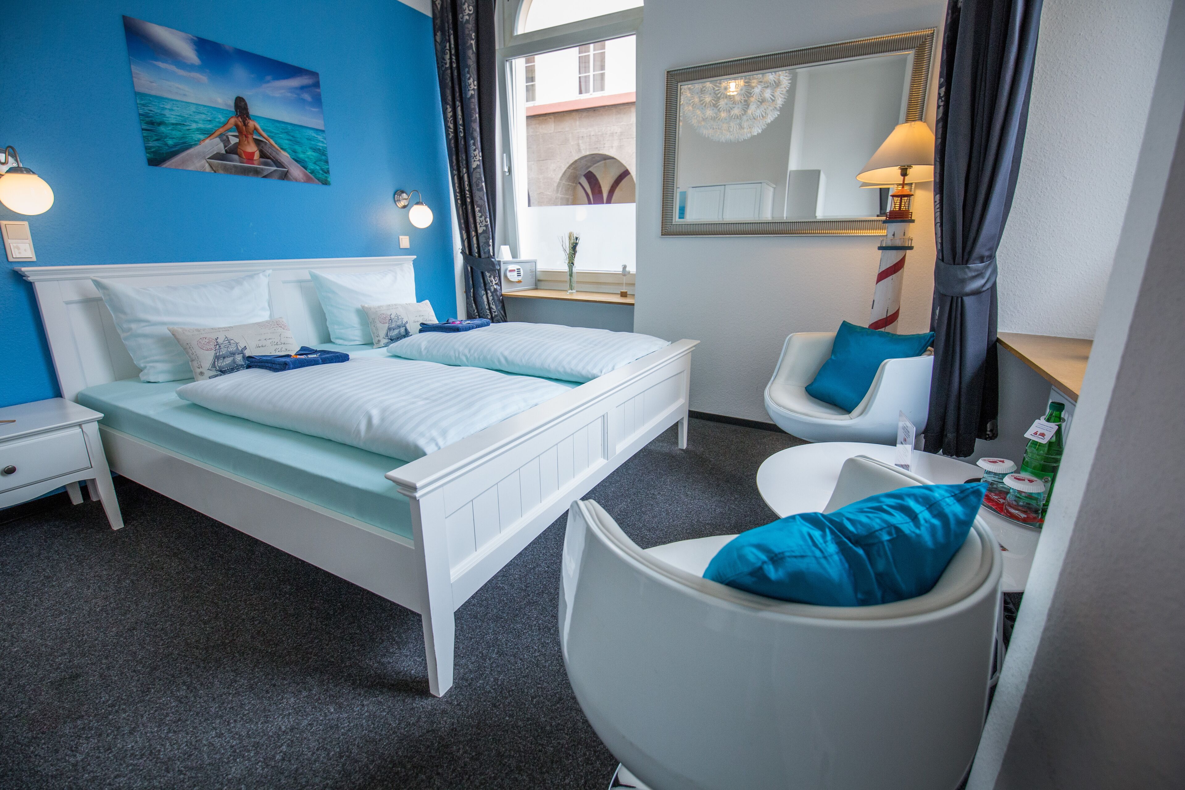 Quarto de hotel com uma cama, uma cadeira, uma escrivaninha, um espelho e uma janela com vista para edifícios.