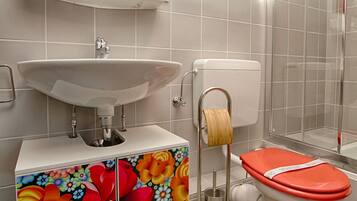 Ein Badezimmer mit weißem Waschbecken, einer Toilette mit rotem Sitz, einer Duschkabine und einem Spiegel über dem Waschbecken.