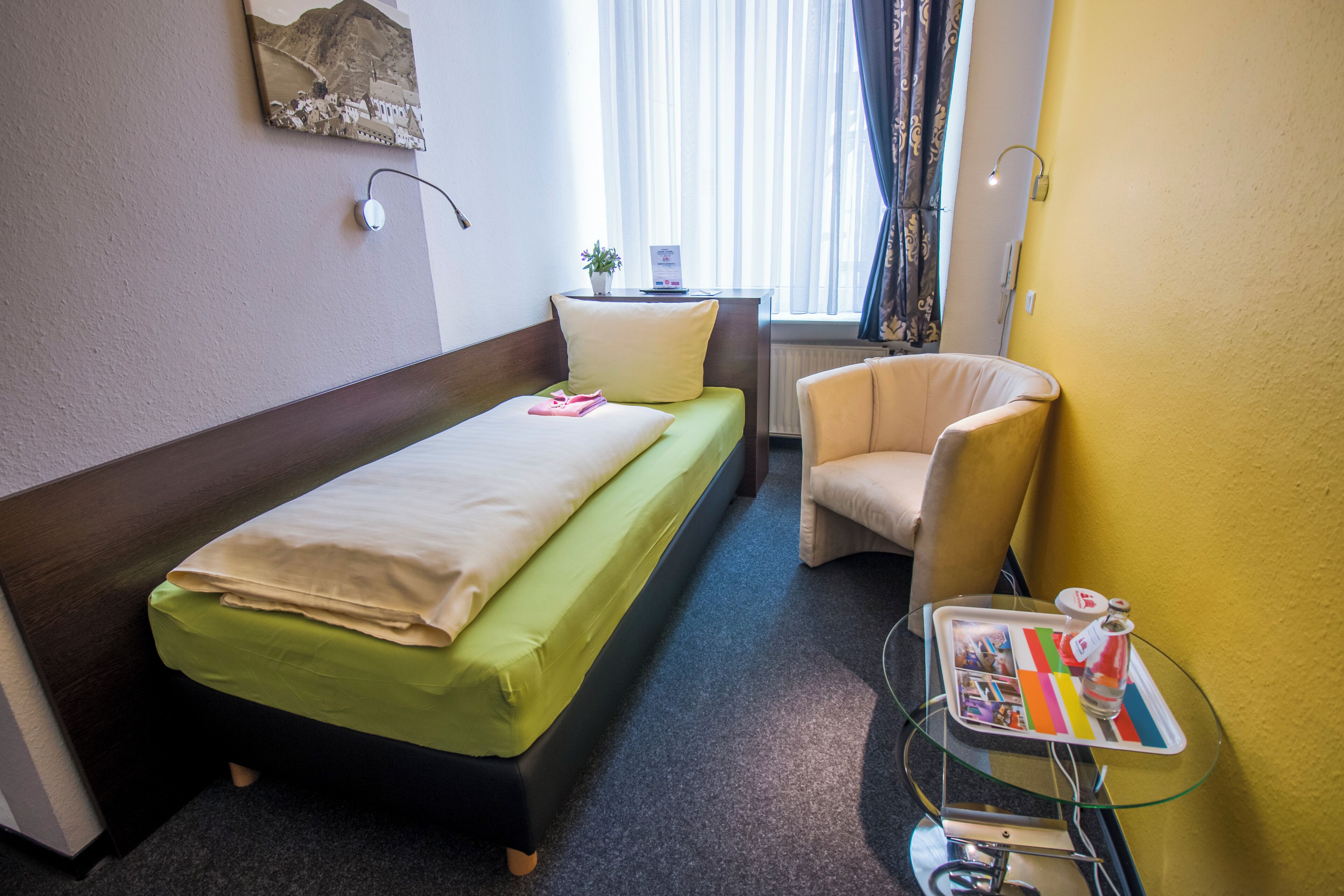A hotel room with a single bed, a chair, a small table with a tray, and a window with curtains.