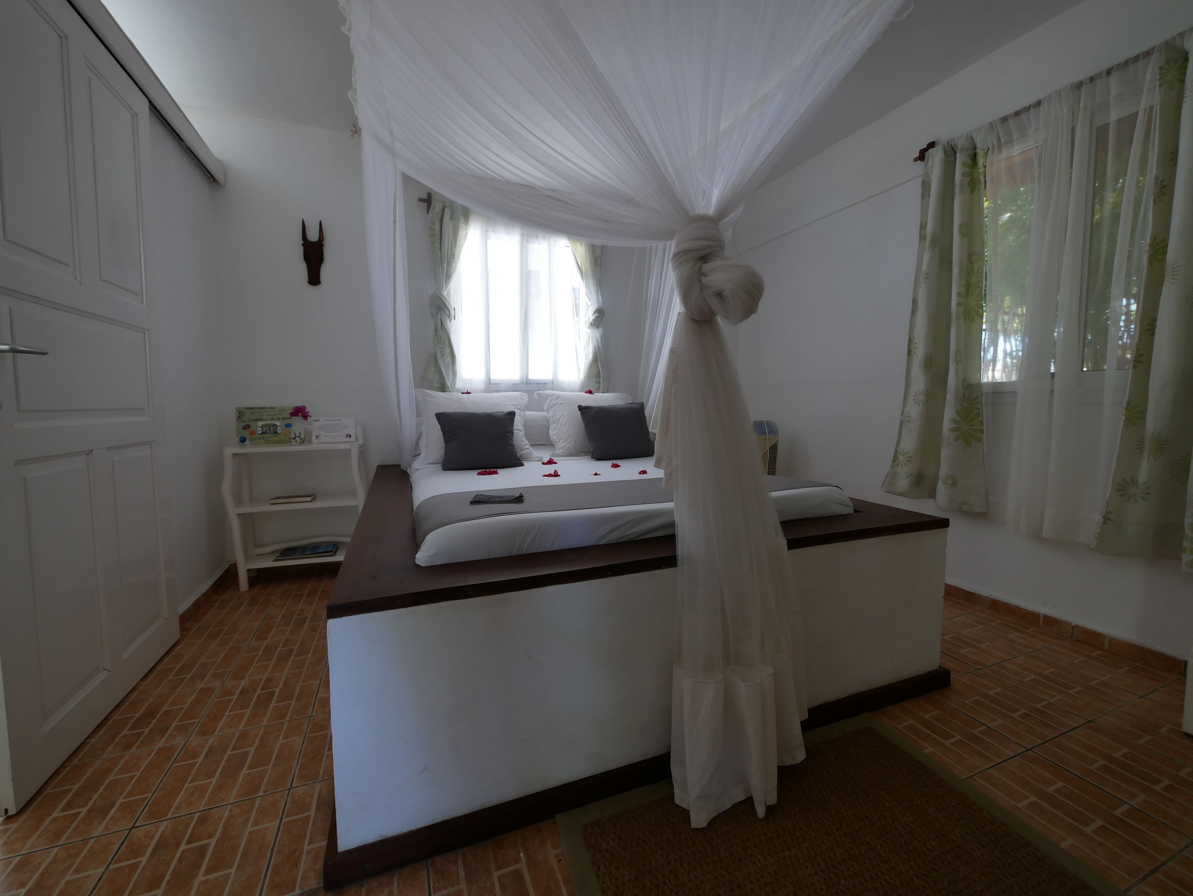 A bedroom with a bed, white curtains, a nightstand, and a wall-mounted clock.