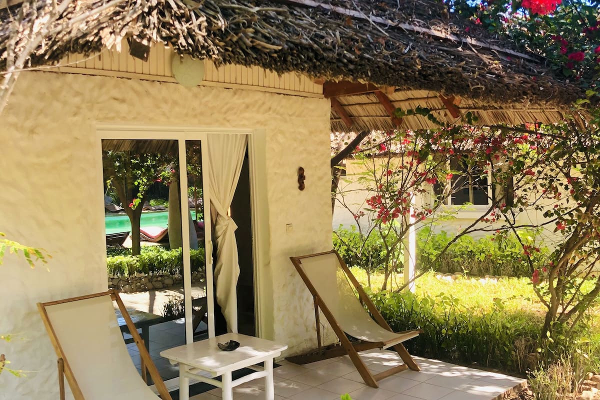 Uma cabana com telhado de palha e uma mesa branca com duas cadeiras de madeira na parte externa.