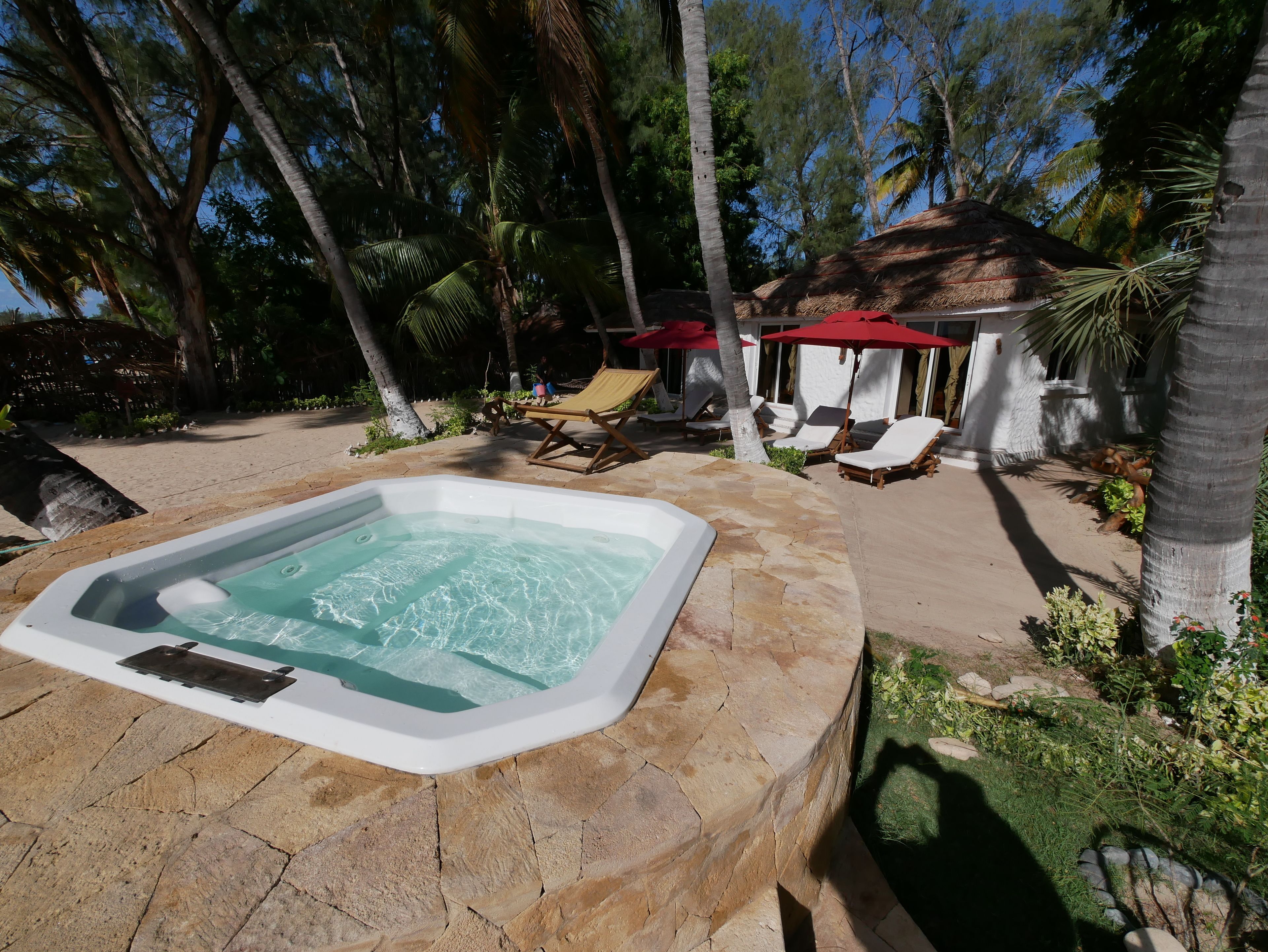 Un jacuzzi rodeado de baldosas de piedra, sillas de descanso y sombrillas en un entorno tropical.
